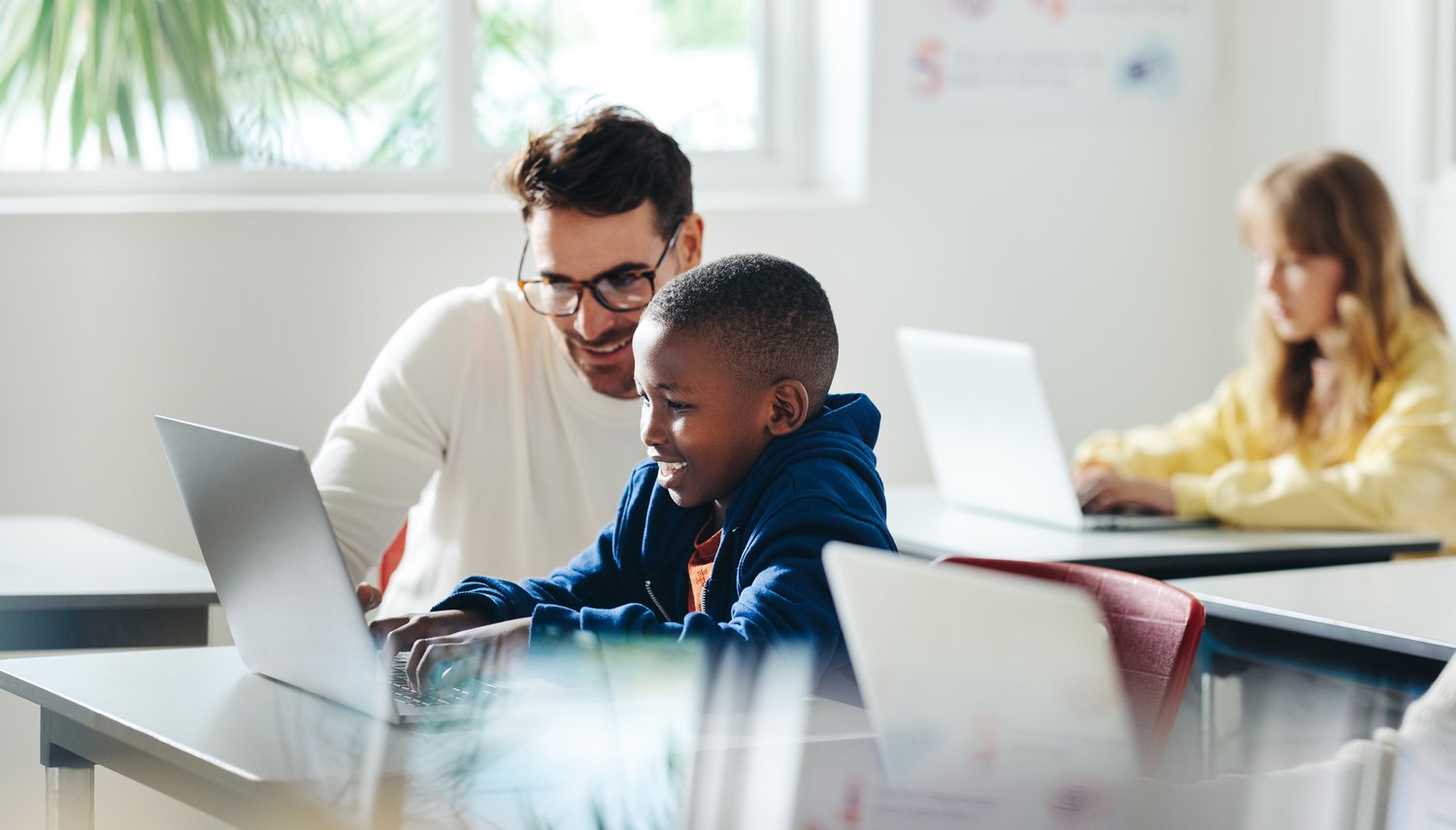 This is an image of a student working on a laptop in class and a teacher is kneeling nearby for support.